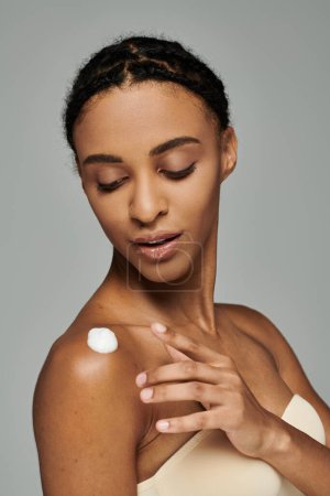 A beautiful young African American woman in a strapless top applying a thick layer of cream to her face on a grey background.