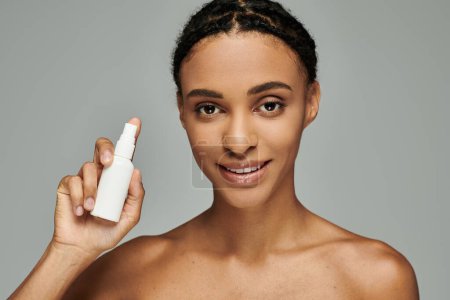 Joven mujer afroamericana en la parte superior sin tirantes sostiene la botella de loción, priorizando la rutina de cuidado de la piel sobre fondo gris.