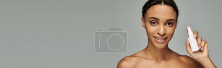 Young African American woman in strapless top gently holding a white skincare product in her right hand against a grey background.