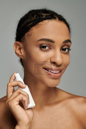 Junge Afroamerikanerin im schulterfreien Top lächelt, während sie ein Kosmetikprodukt auf grauem Hintergrund hält.