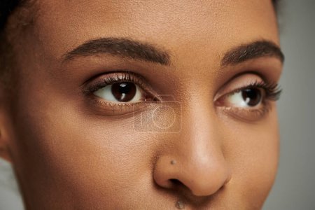 Téléchargez les photos : Gros plan d'une jeune afro-américaine aux yeux marron époustouflants, routine de soins de la peau, sur fond gris. - en image libre de droit