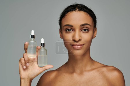 Femme afro-américaine en haut sans bretelles tenant bouteille de produit de soins de la peau sur fond gris.