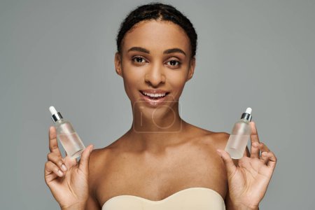 Young African American woman in strapless top taking care of skin, holding two serum bottles on grey background.