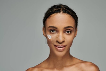 Photo for Young African American woman in strapless top, delicately applying cream to her face on a serene grey background. - Royalty Free Image