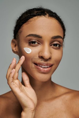 Photo for Young African American woman in strapless top applying cream on her face against a grey backdrop. - Royalty Free Image
