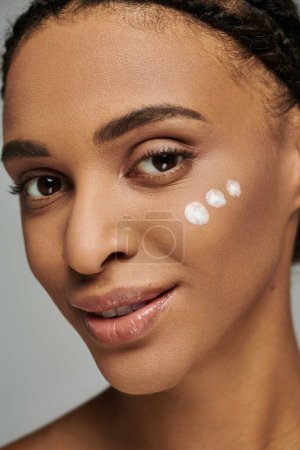 Junge Afroamerikanerin in trägerlosem Top mit weißen Punkten aus Creme im Gesicht, pflegt die Haut vor grauem Hintergrund.