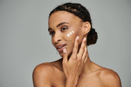 Junge Afroamerikanerin mit cremefarbenem Gesicht, konzentriert auf die Hautpflege, in einem schulterfreien Oberteil vor grauem Hintergrund.