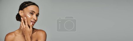 Photo for Young African American woman in strapless top touching her face gently, caring for her skin, on grey background. - Royalty Free Image