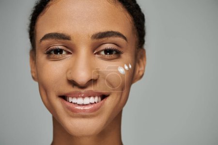 Foto de Joven mujer afroamericana en top sin tirantes, brillando con maquillaje, sonriendo sobre fondo gris. - Imagen libre de derechos