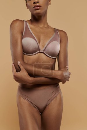 Foto de Mujer afroamericana en sujetador y bragas posa con gracia sobre un fondo beige, mostrando su confianza y rutina de cuidado corporal. - Imagen libre de derechos