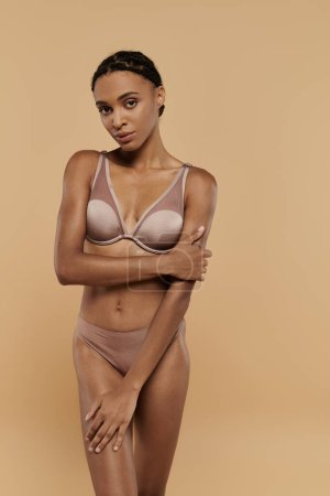 A pretty and slim African American woman in nude bra and panties, gracefully tending her body on a beige background.