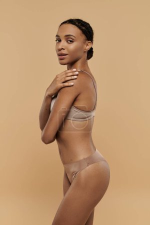 Foto de Una mujer afroamericana bonita y delgada en bikini bronceado posa elegantemente para la cámara sobre un fondo beige suave. - Imagen libre de derechos