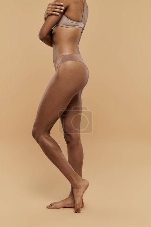 An African American woman showcases her toned body in bikini and panties on a beige background.