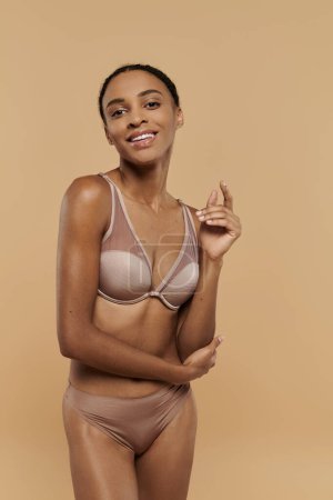 Eine atemberaubende, schlanke Afroamerikanerin posiert elegant in einem nackten BH, der auf einem weichen beigen Hintergrund Selbstvertrauen und Selbstbewusstsein ausstrahlt..