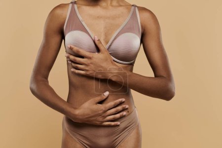 Téléchargez les photos : Femme afro-américaine en bikini bronzé tenant sereinement son estomac sur fond beige. - en image libre de droit