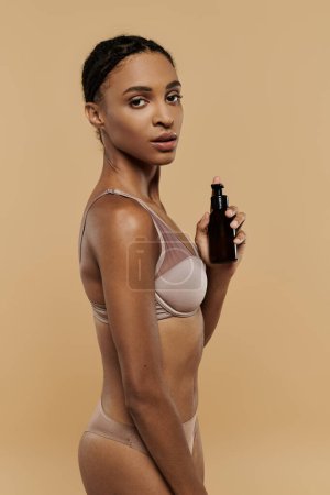 A slim African American woman in a bikini enjoys a bottle of body oil against a beige backdrop.