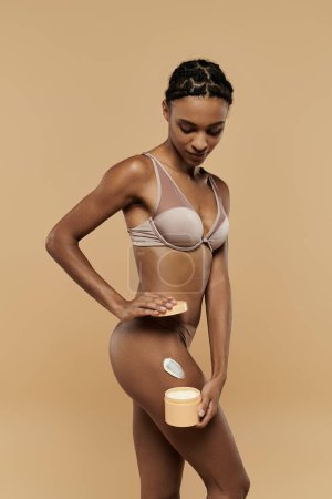 Foto de Una impresionante mujer afroamericana en bikini sostiene delicadamente un frasco de crema sobre un fondo beige. - Imagen libre de derechos