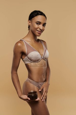 Slim African American woman in bikini holding sugar scrub against a beige backdrop.