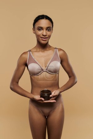 Una esbelta mujer afroamericana en bikini sosteniendo un exfoliante de café sobre un fondo beige.