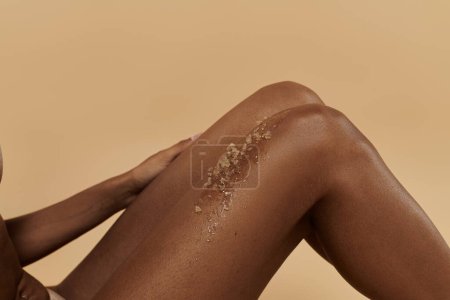slim African American woman with coffee scrub on legs on a beige background.