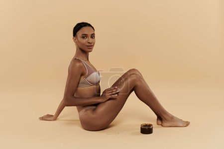 Photo for A pretty, slim African American woman in a bikini is sitting on the ground, taking care of her body in a serene setting. - Royalty Free Image