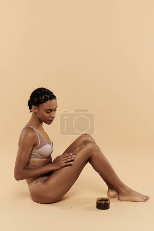 Slim African American woman in underwear sits gracefully with coffee scrub on legs on a beige background.