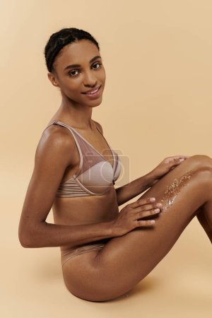 A slim African American woman seated gracefully with coffee scrub on legs on a beige background.