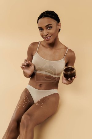 Mujer afroamericana delgada en bikini con exfoliante de azúcar en las piernas sobre fondo beige.