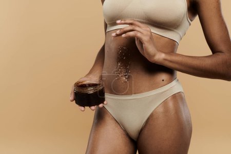 Una impresionante mujer afroamericana en bikini seductora sostiene un frasco de exfoliante sobre un fondo beige.