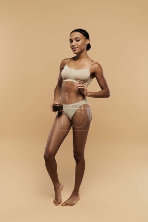 An elegant African American woman showcases her toned figure in a bikini against a beige backdrop.