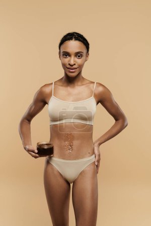 Mujer afroamericana en bikini con elegancia sosteniendo un exfoliante sobre un fondo beige.