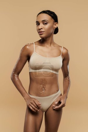 A pretty, slim African American woman is seen taking care of her body in tan lingerie on a beige background.