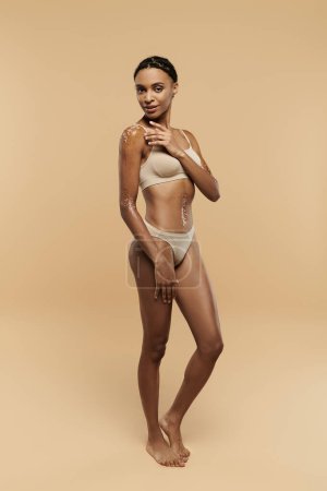 A stunning African American woman confidently poses in a bikini on a beige background.
