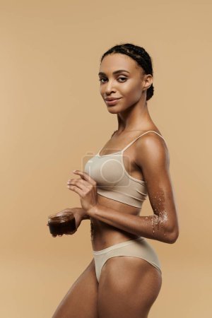 Foto de Una hermosa mujer afroamericana en bikini sosteniendo un exfoliante sobre un fondo beige. - Imagen libre de derechos