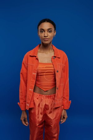 Photo for A stunning young African American woman stands confidently in front of a vibrant blue background, wearing a striking orange outfit. - Royalty Free Image