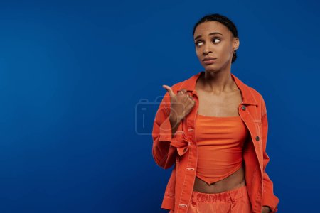 Young African American woman confidently points to something while wearing a vibrant orange top on a blue background.