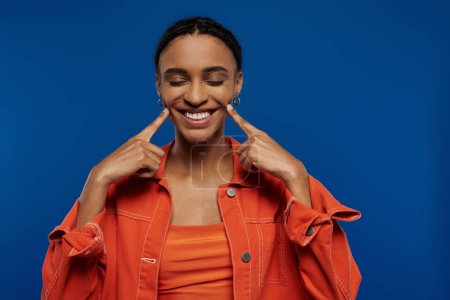 A pretty young African American woman in a vibrant orange shirt on a blue background making a funny face.
