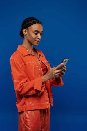 Young African American woman in bright orange outfit engrossed in cell phone on a blue background.