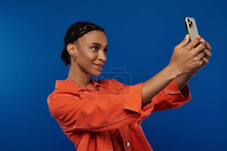 Photo for A young African American woman in vibrant orange outfit taking a picture with her cell phone against a blue background. - Royalty Free Image