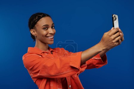 Foto de Vibrant young African American woman in orange outfit taking a photo with her cell phone against a vivid blue background. - Imagen libre de derechos