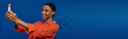Vibrant young African American woman in orange outfit taking a picture with her cell phone against a bold blue backdrop.