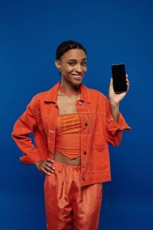 Una joven y elegante mujer afroamericana con un traje naranja brillante, sosteniendo un teléfono celular sobre un fondo azul.