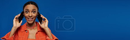 Young African American woman in vibrant orange outfit holding two cell phones to her ears on a blue background.