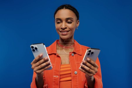 Foto de Una joven afroamericana con un vibrante atuendo naranja sostiene dos teléfonos celulares en sus manos sobre un fondo azul. - Imagen libre de derechos