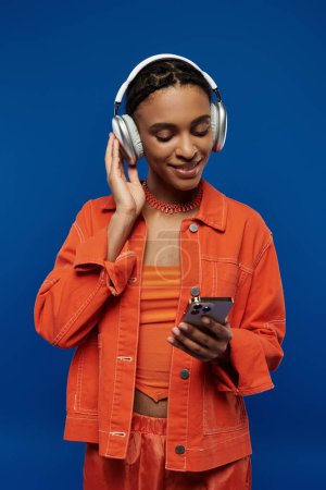 Junge Afroamerikanerin in orangefarbenem Outfit, Kopfhörer tragend, in Handybildschirm auf blauem Hintergrund versunken.