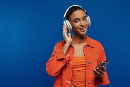 Joven mujer afroamericana en traje naranja vibrante escuchando música en los auriculares mientras sostiene un teléfono celular.