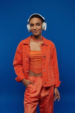 A stylish African American woman in orange stands with headphones against a vibrant blue background.