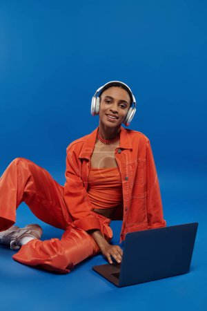 Vibrant young African American woman in orange outfit sits on floor, absorbed in laptop with headphones.