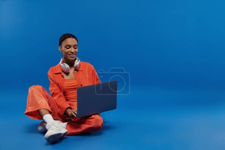 Téléchargez les photos : Une jeune afro-américaine dans une tenue orange vibrante s'assoit sur le sol avec un ordinateur portable devant elle. - en image libre de droit