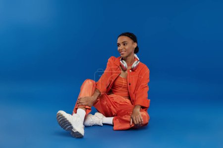 Téléchargez les photos : Une jeune Afro-Américaine dans une tenue orange frappante assise gracieusement sur le sol sur un fond bleu vif. - en image libre de droit